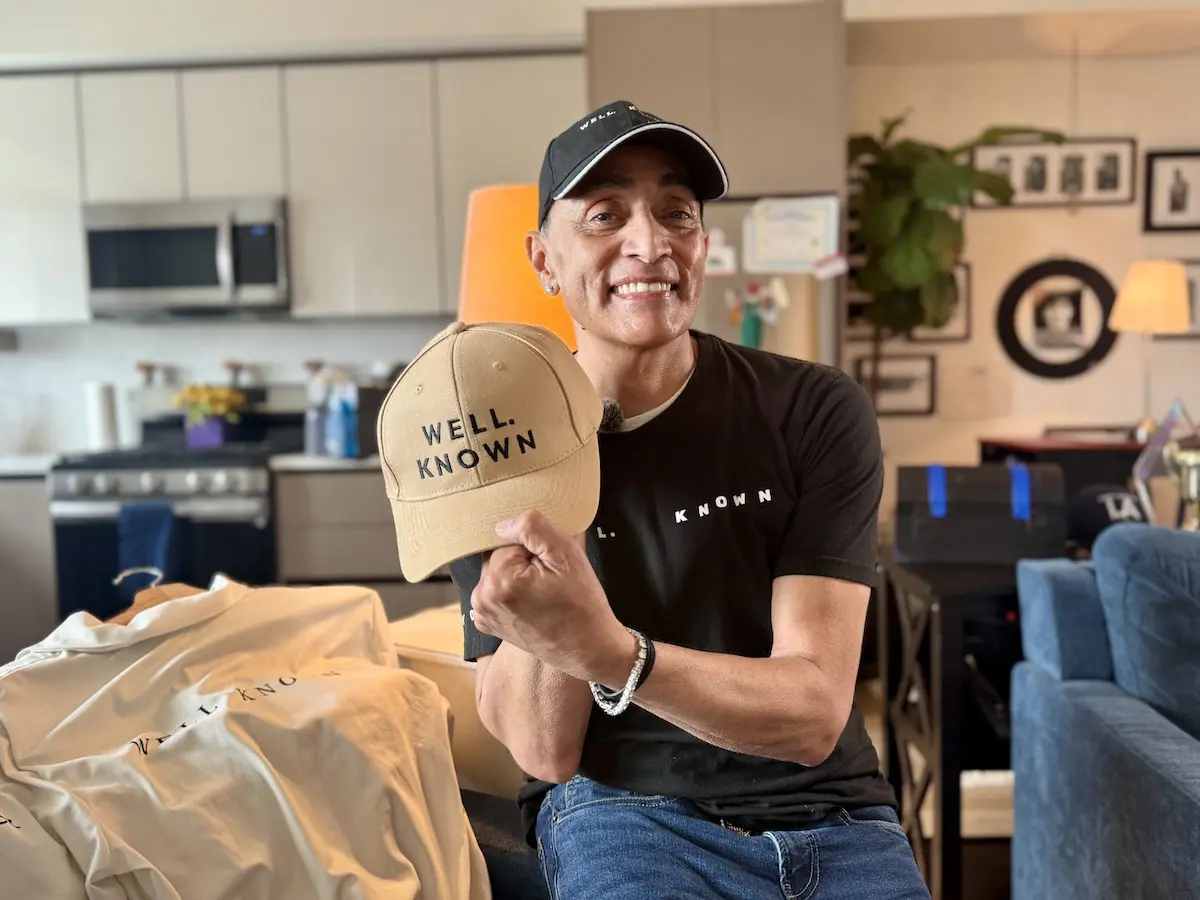 Daniel Castilleja smiles while holding a tan ‘WELL. KNOWN’ cap from his clothing brand, photographed in his apartment