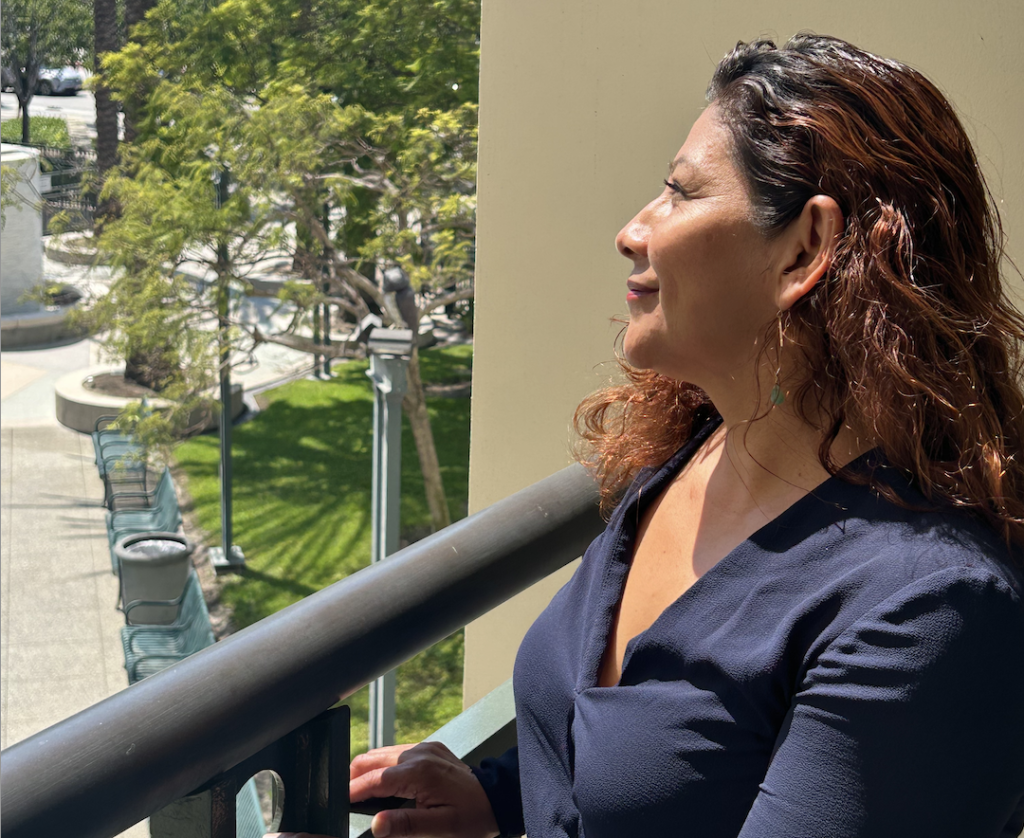 Elizabeth Rodriguez stands on a balcony in Culver City, looking out toward palm trees and sunlight, reflecting on her journey with St. Joseph Center.