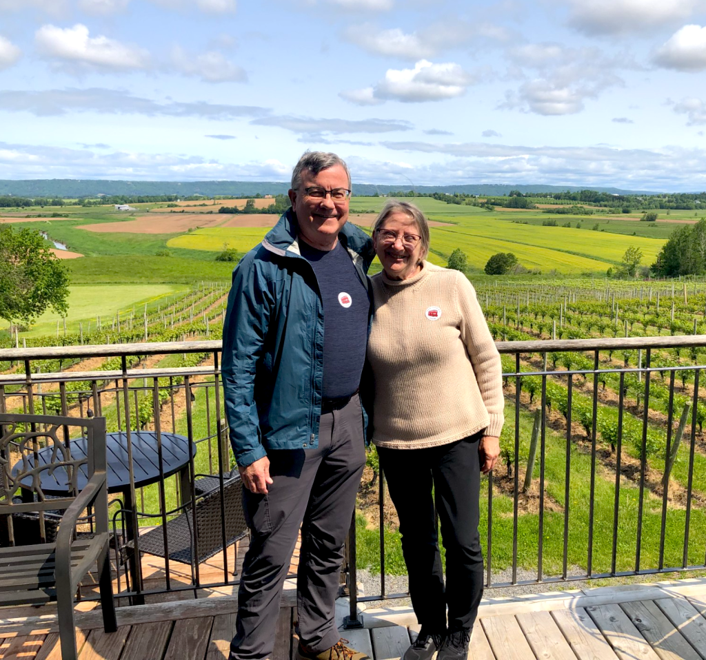 Hope and Steve Heaney stand together on a sunny terrace overlooking rolling green farmland, smiling warmly at the camera.