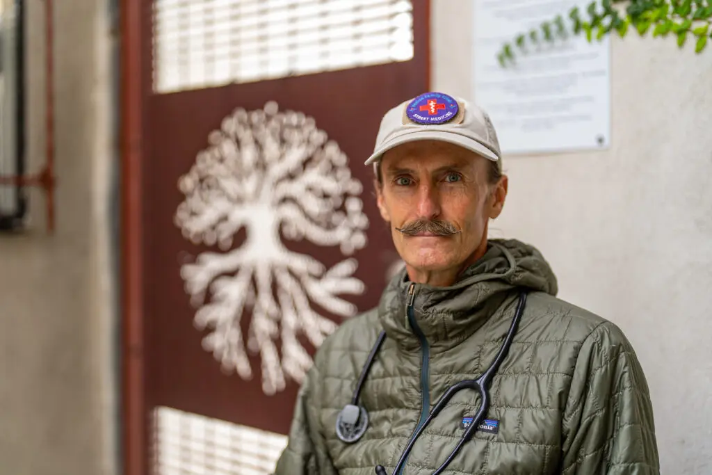 Dr. Coley King smiling at the camera in a baseball cap and green jacket, standing in front of a St. Joseph Center backdrop.