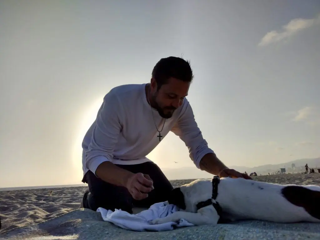 Derrick Rutledge leaning forward on the beach, wearing a white long-sleeved shirt with the ocean and bright sky behind him.