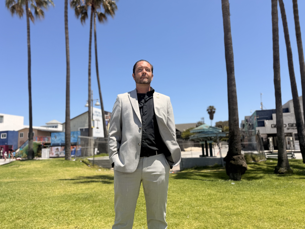 Derrick Rutledge standing in a light-colored suit and black shirt on a grassy area near Venice Beach, with palm trees in the background.