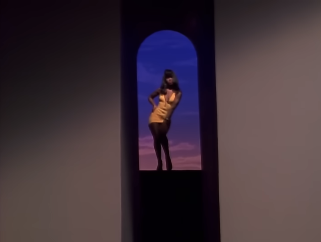 A dancer wearing a gold outfit poses against a purple backdrop in a still from Sir Mix-a-Lot’s “Baby Got Back” music video.
