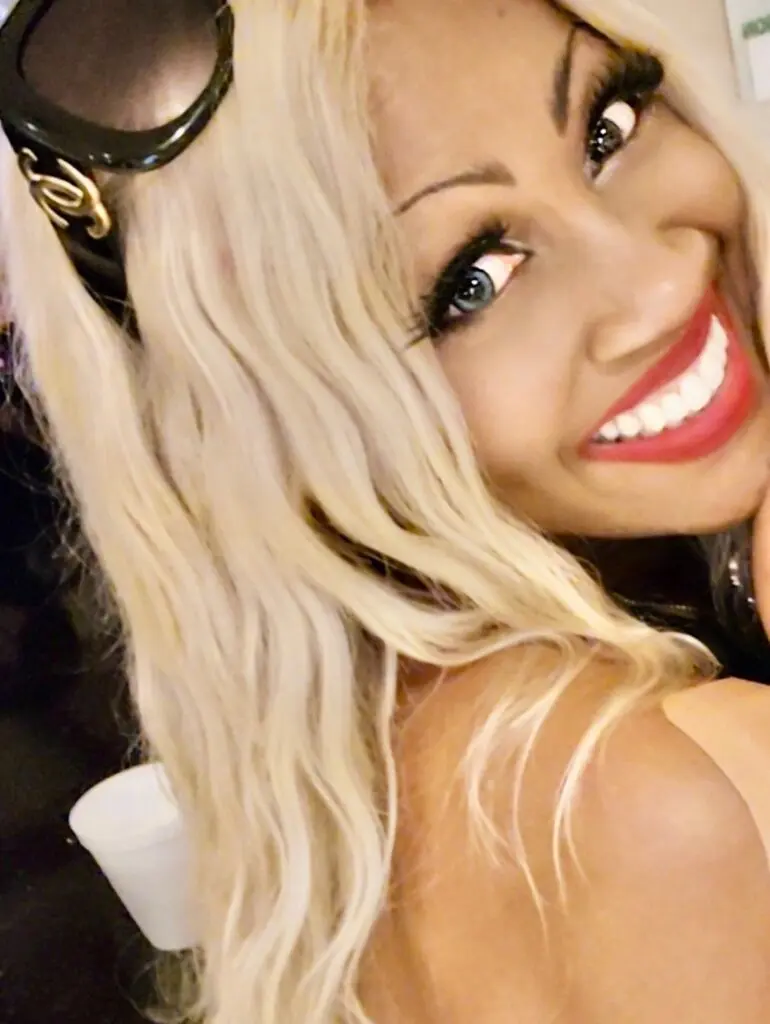 Shuren Joice smiling at the camera with long blonde hair and sunglasses resting on her head.