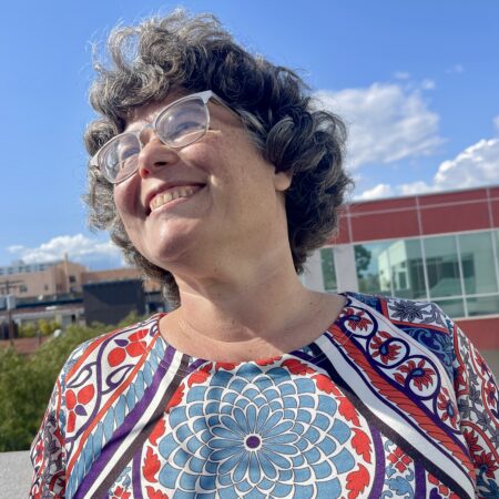 Chef Sarah, smiling brightly with curly salt-and-pepper hair and clear glasses, gazes upward under a blue sky. She’s wearing a colorful, patterned top in red, blue, and purple.