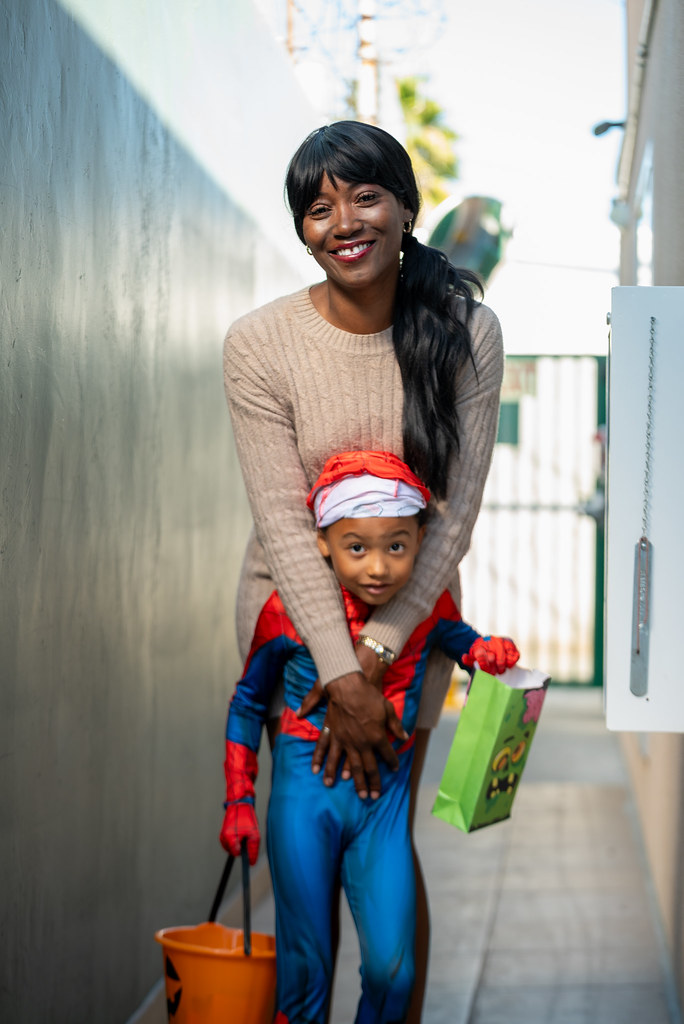 Cheraya hugging her young son, who is dressed in a Spider-Man costume and holding a pumpkin bucket, as they stand together in an outdoor walkway.