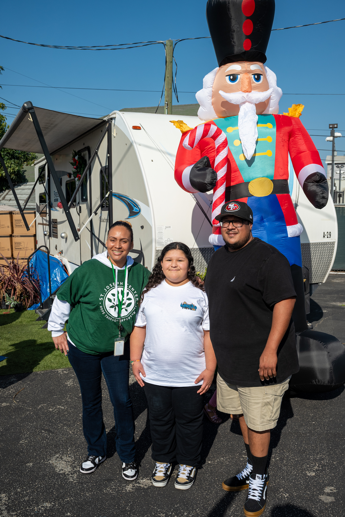 Charles and his daughter Irie standing with a St. Joseph Center staff member at Client Appreciation Day, smiling outdoors with a large holiday nutcracker decoration behind them.