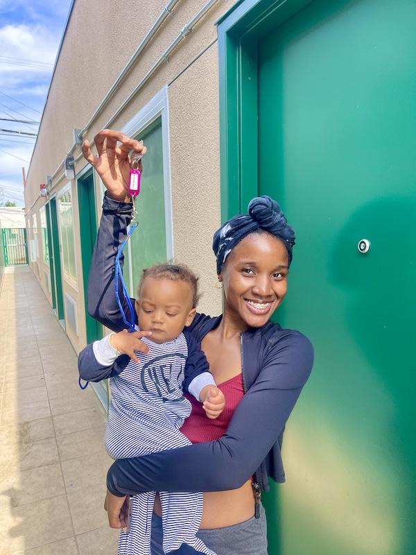 Nandi smiles while holding her child and raising a set of keys outside La Ramona Housing Center, marking a milestone in her journey to stable housing.