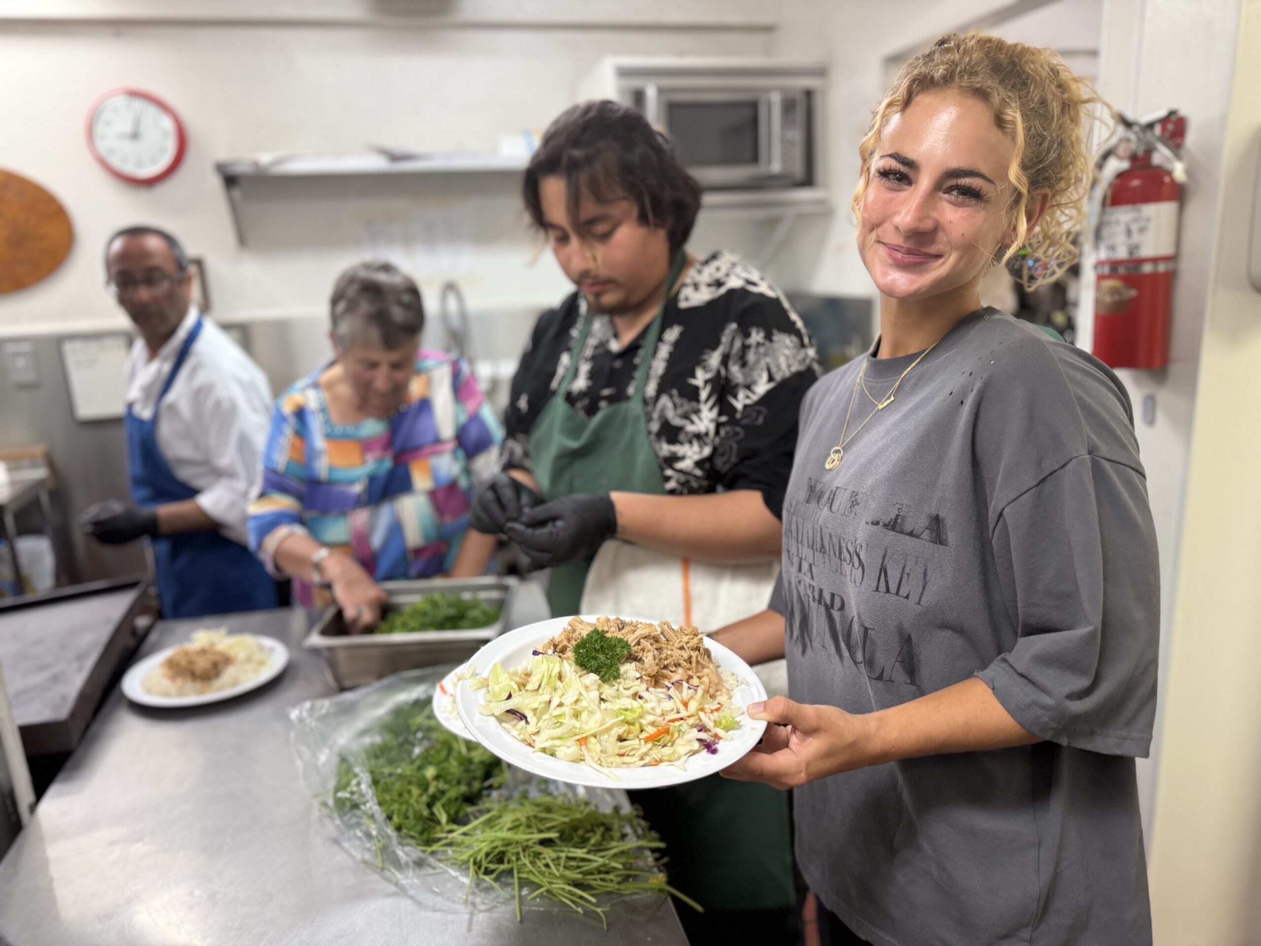 Claire smiling while holding a plate of food in the Bread and Roses Café kitchen as volunteers prepare meals in the background