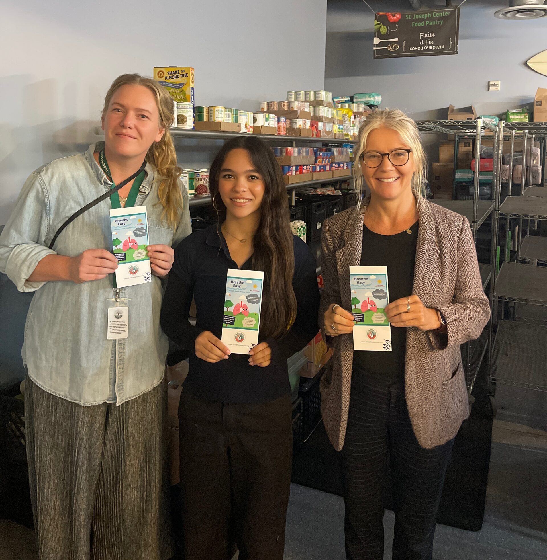 Sienna stands with St. Joseph Center staff holding Breathe Easy educational materials inside the Bread and Roses Food Pantry