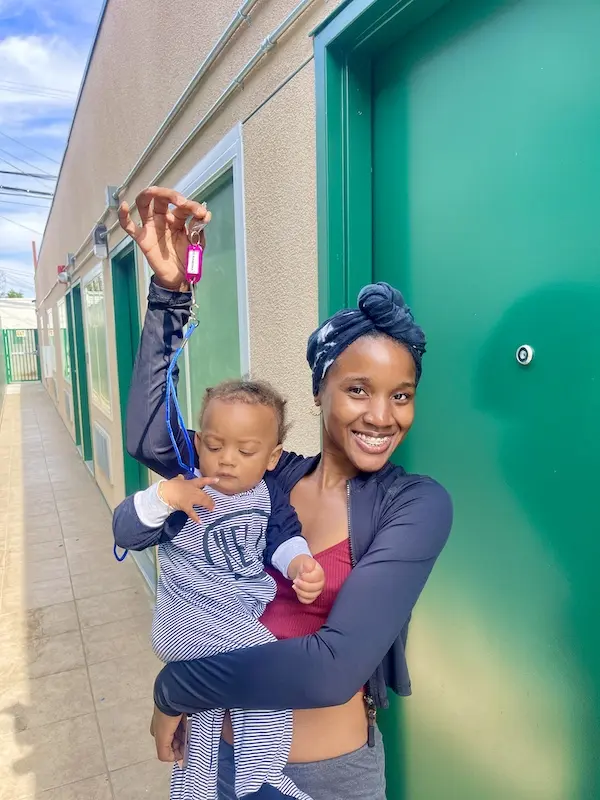 Nandi smiles while holding her child and raising a set of keys outside La Ramona Housing Center, marking a milestone in her journey to stable housing.