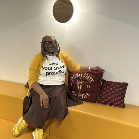 Suzette Shaw sits smiling on a modern yellow bench at ASU Local, wearing a “Wear Your Crown Proudly” shirt, yellow sneakers, and a gold cardigan. She’s seated next to Arizona State University pillows, representing her return to student life.